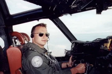 A young Brian Vinci in the cockpit of a P-3 Orion wearing a flight suit, headset, and aviator sunglasses